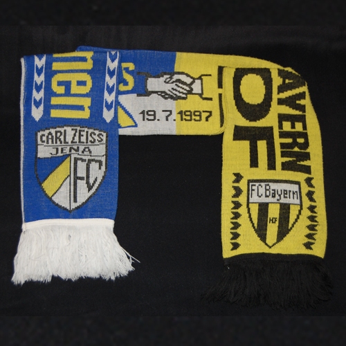 Unter dem Motto „Eine Region steht zusammen“ empfing der FC Bayern Hof am 19. Juli 1997 den FC Carl Zeiss Jena, der damals der 2. Bundesliga angehörte. 3000 Zuschauer bestaunten einen Freistoßtreffer des 23-jährigen Bernd Schneider, der auf der Grünen Au schon einmal andeutete, warum er es bis zum Vizeweltmeister bringen sollte. Insgesamt fielen sieben Tore, leider aber keines für Gelbschwarz. Neben diesem Schal (Leihgabe von Jörg Herr), der zum Freundschaftsspiel aufgelegt wurde, kannst Du Dir hier im Museum auch den Fernsehspielbericht ansehen.